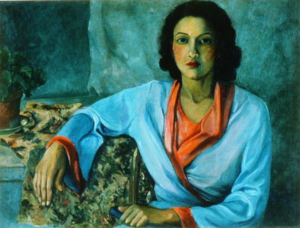 A portrait of a black woman, seated, one hand in her lap, one resting on a table. She is wearing a light blue with red trim long sleeved top. 