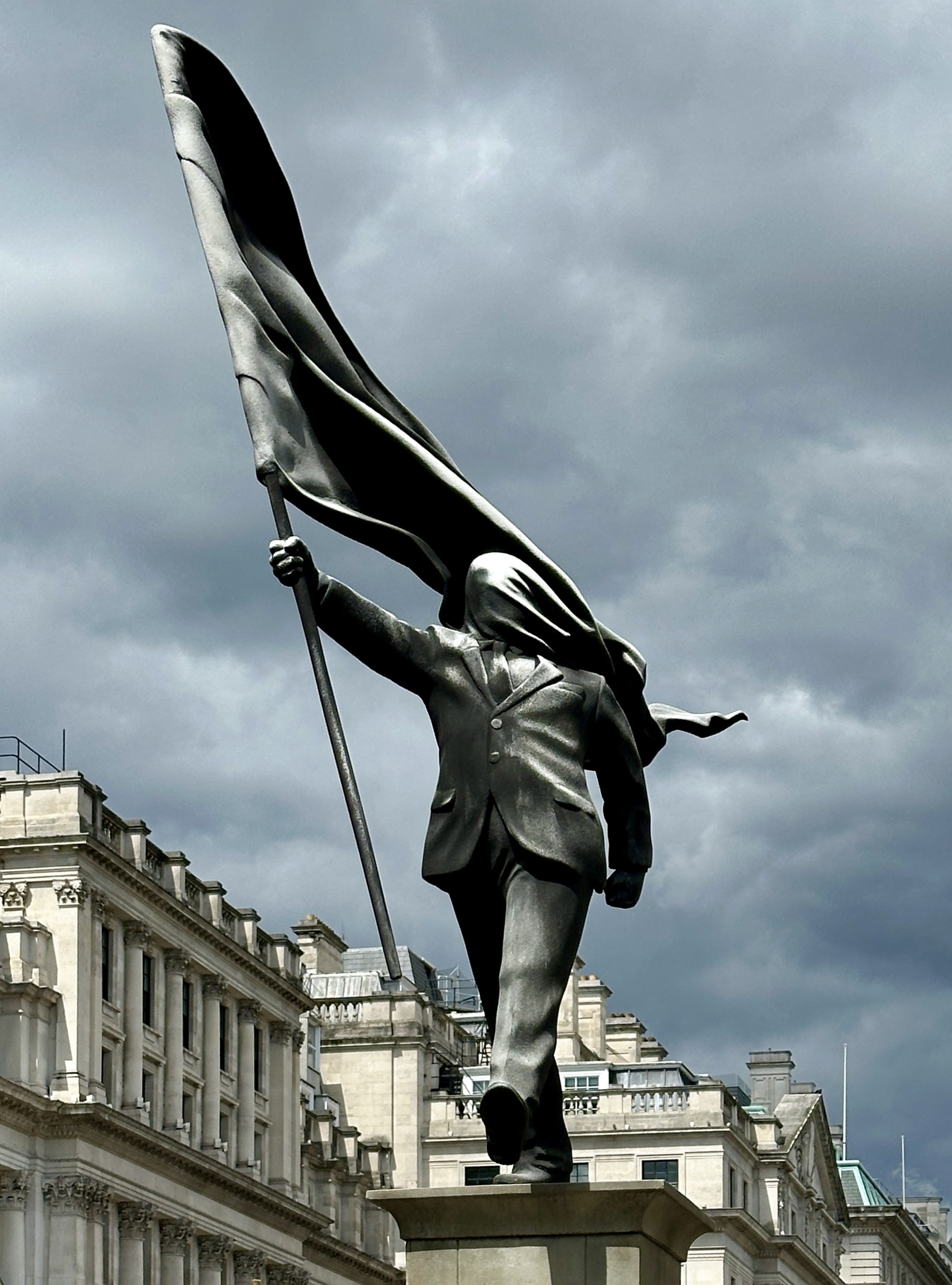 A statue of a man, walking, carrying a flag which has wrapped around his face, making it impossible for him to see where he is going. He is about to stepping off the edge of a high plinth